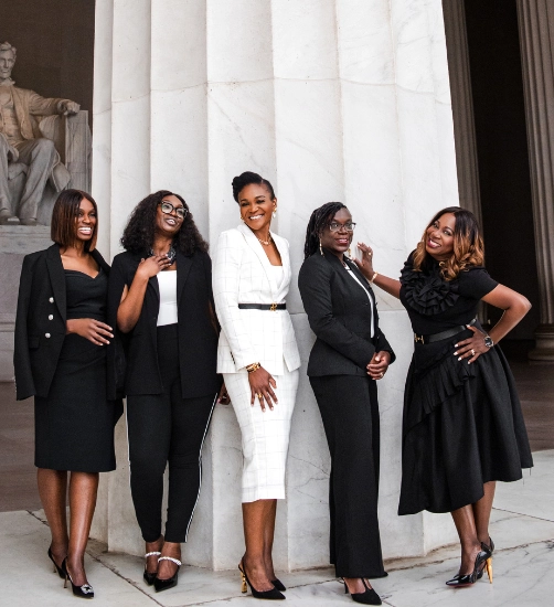 Five stylish women pose confidently in formal black and white outfits, standing in front of a large white column at the Lincoln Memorial, with the Lincoln statue visible in the background.
